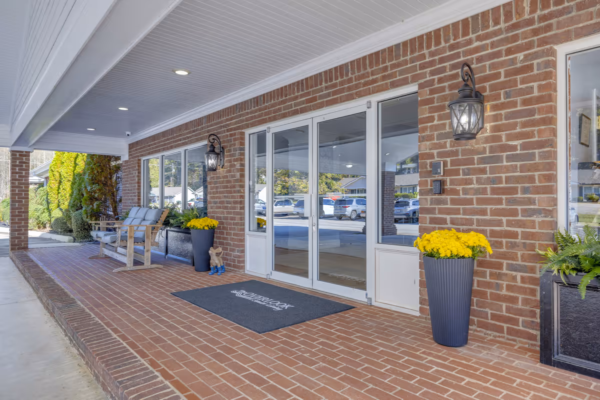 Covered brick entrance of a building with glass double doors, two black wall lanterns, potted yellow flowers, a bench with cushions, and a welcome mat that reads 'Overlook'. Cars and greenery are visible in the background.