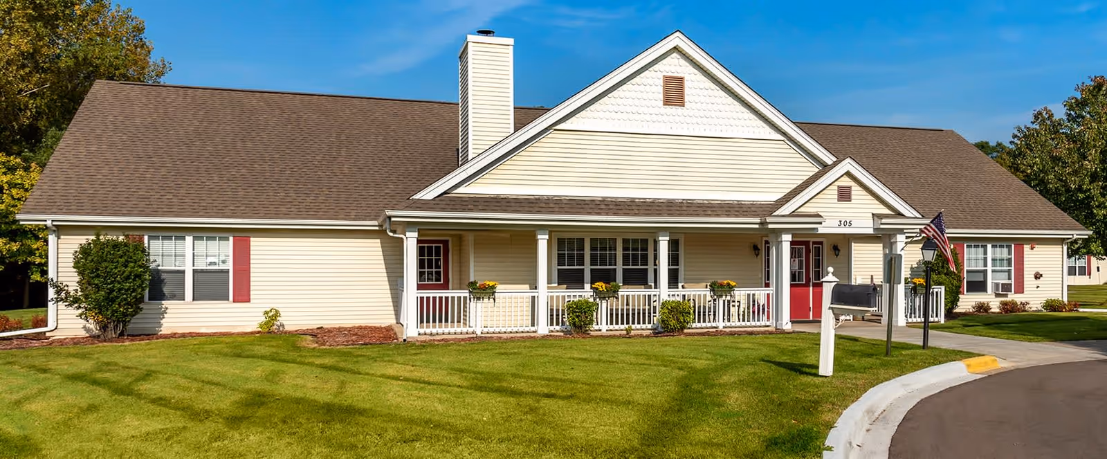 Single-story residential building with beige siding, brown roof, and white trim. The building has a front porch with white railings and several windows with red shutters. There is a well-maintained green lawn in front, a mailbox, and an American flag near the entrance. The sky is clear and blue.