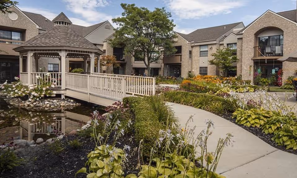 Landscaped courtyard with a gazebo and footbridge over a pond in front of a multi-story residential building.