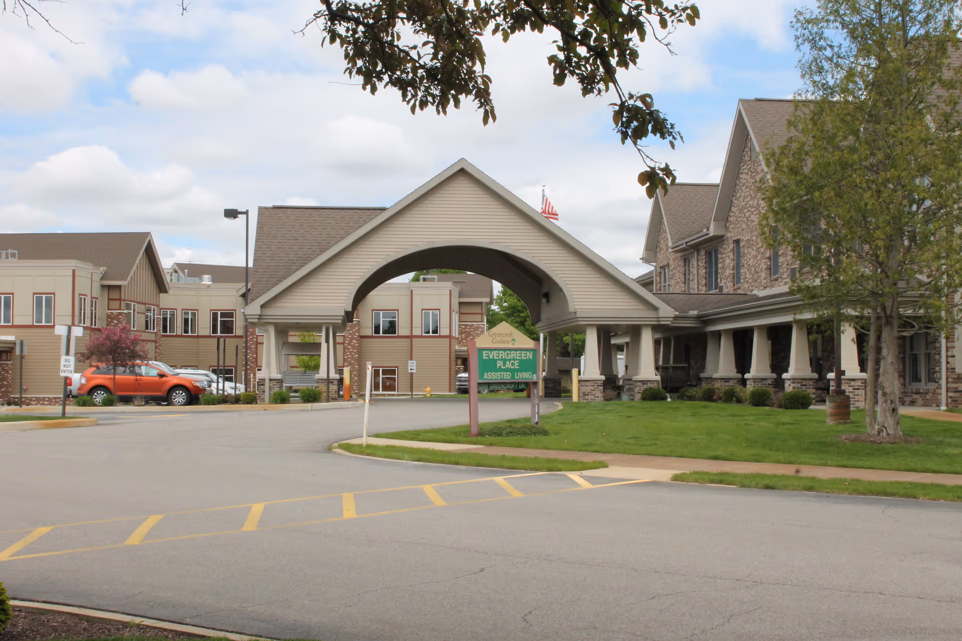 Exterior view of Evergreen Place assisted living facility showing the main entrance with a covered driveway, a green sign with the facility name, a parking area with a red vehicle, and surrounding buildings with stone and siding exteriors under a partly cloudy sky.
