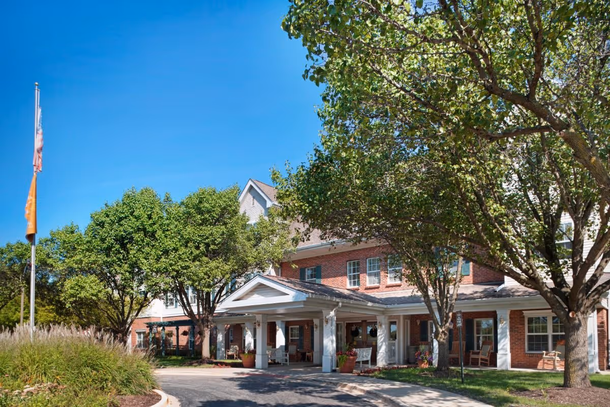 Exterior view of Brighton Gardens of Wheaton senior living facility showing a brick building with white trim, a covered entrance with columns, surrounded by green trees and a clear blue sky. There are flagpoles with flags on the left side and a driveway leading to the entrance.