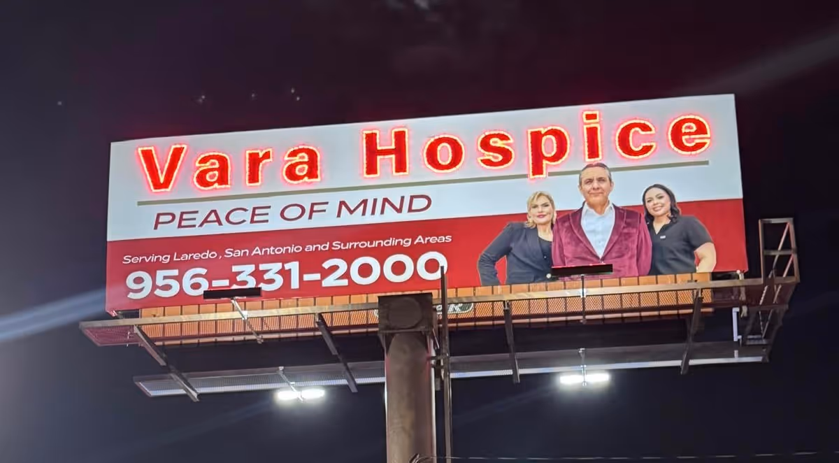 Nighttime outdoor billboard advertising Vara Hospice with three people displayed.