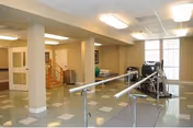 Rehabilitation room with parallel bars, exercise equipment, and large windows in a care facility.