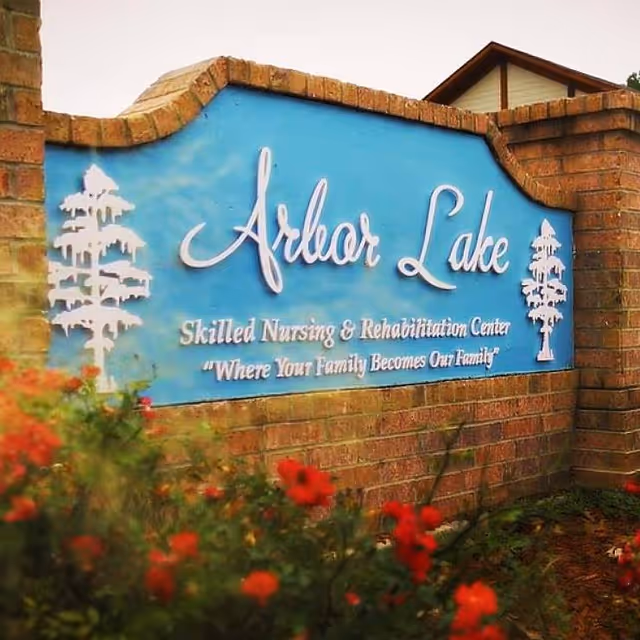 A brick sign for Arbor Lake Skilled Nursing & Rehabilitation Center with the slogan 'Where Your Family Becomes Our Family' on a blue background, surrounded by red flowers and greenery.