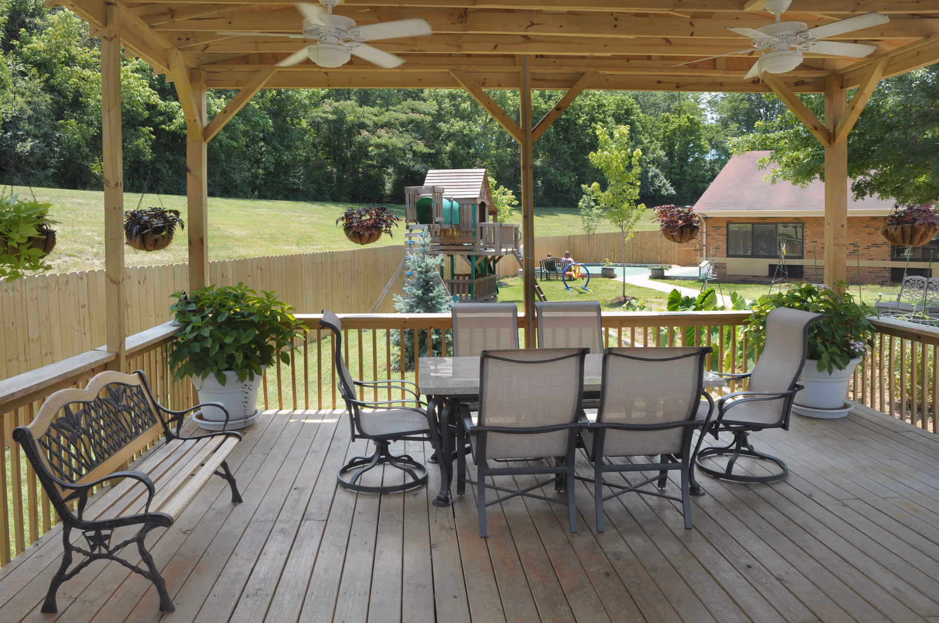 Covered wooden patio with a dining table and six chairs, two ceiling fans, hanging flower pots, potted plants, a bench, and a view of a fenced grassy yard with a playground and a brick building in the background.
