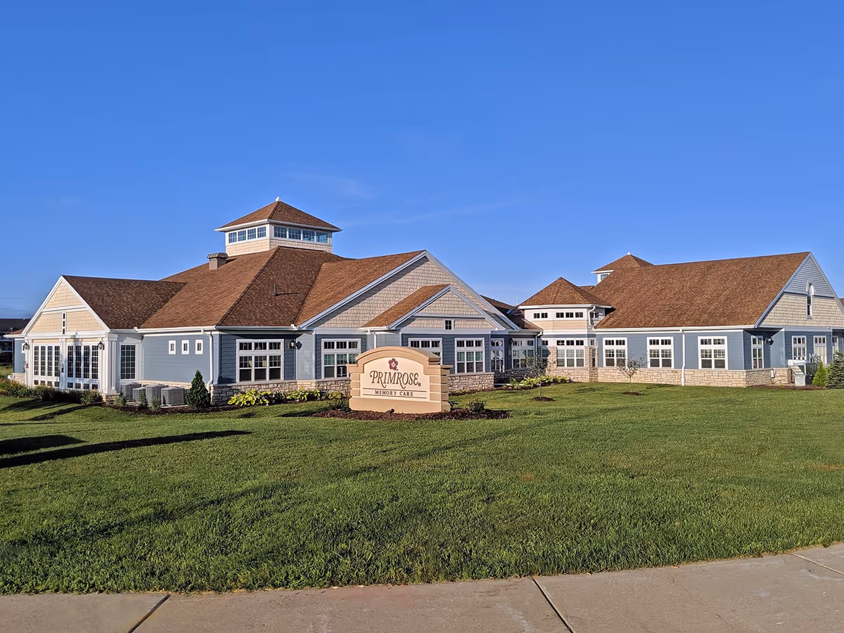 Exterior view of Primrose Retirement Community of Wausau building with blue siding, brown shingled roofs, multiple windows, and a sign in front that reads 'Primrose Memory Care'. The building is surrounded by a well-maintained green lawn under a clear blue sky.