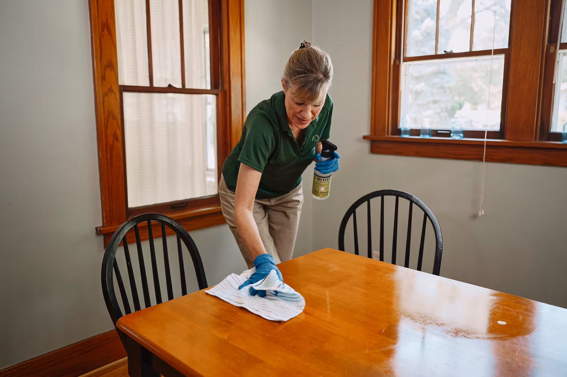 Caregiver in a green shirt cleans a wooden dining table indoors.