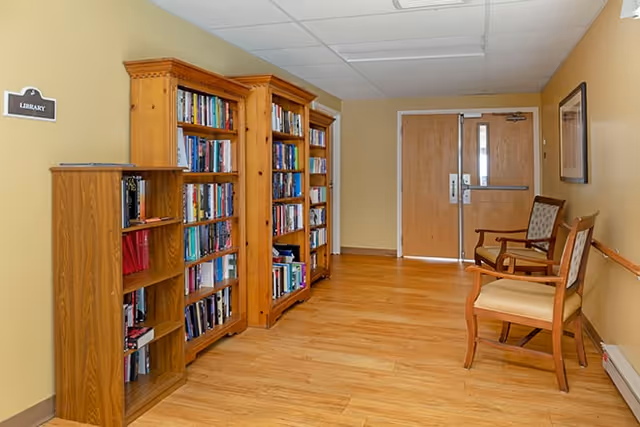 Interior hallway area with wooden bookshelves filled with books on the left side, a sign labeled 'Library' above the shelves, two wooden chairs with cushioned seats on the right side, beige walls, and double wooden doors at the end of the hallway.