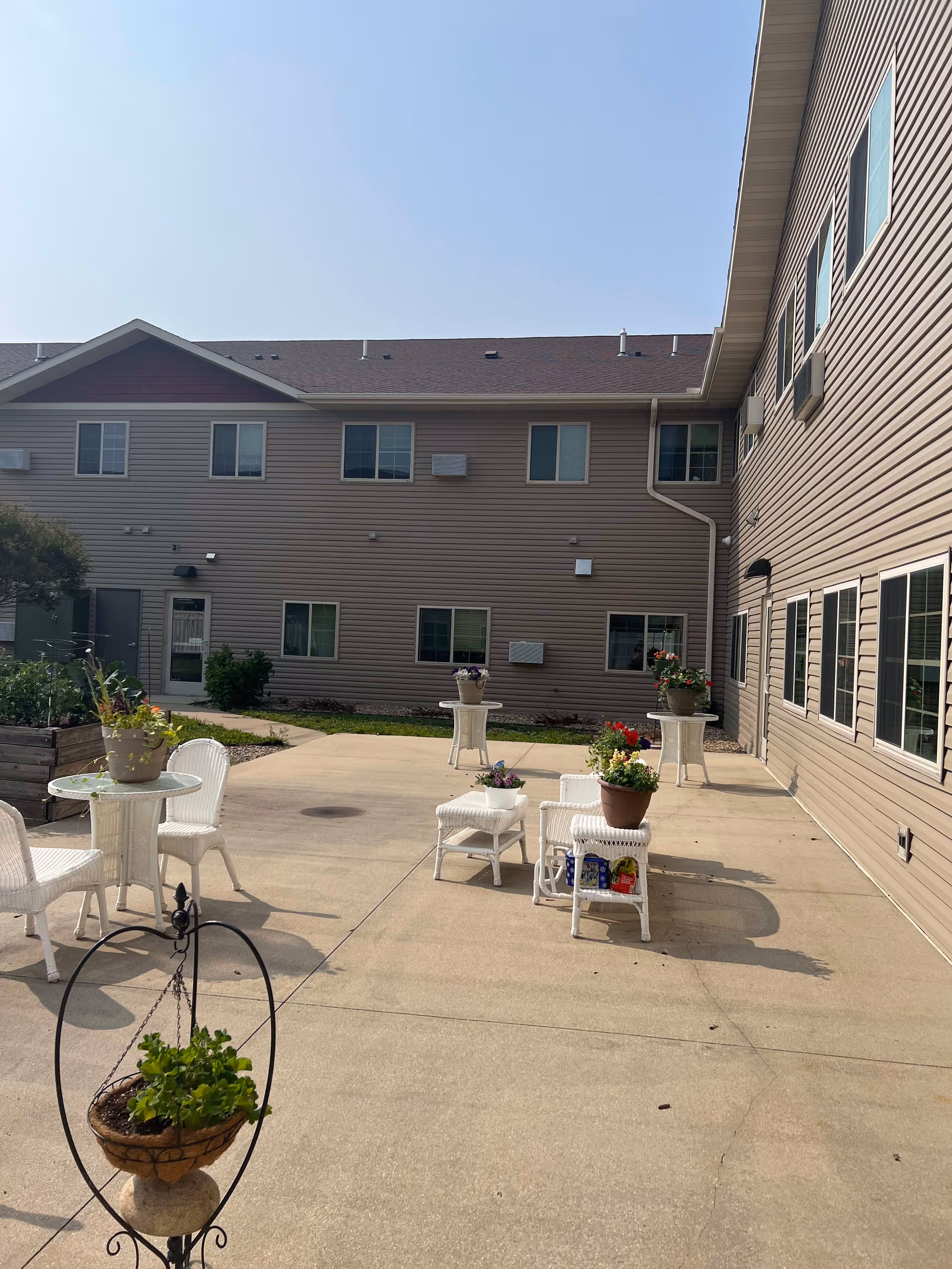 Outdoor courtyard area of a senior living facility with beige siding buildings surrounding a concrete patio. The patio has white wicker chairs and tables, some with potted plants and flowers. The sky is clear and blue.