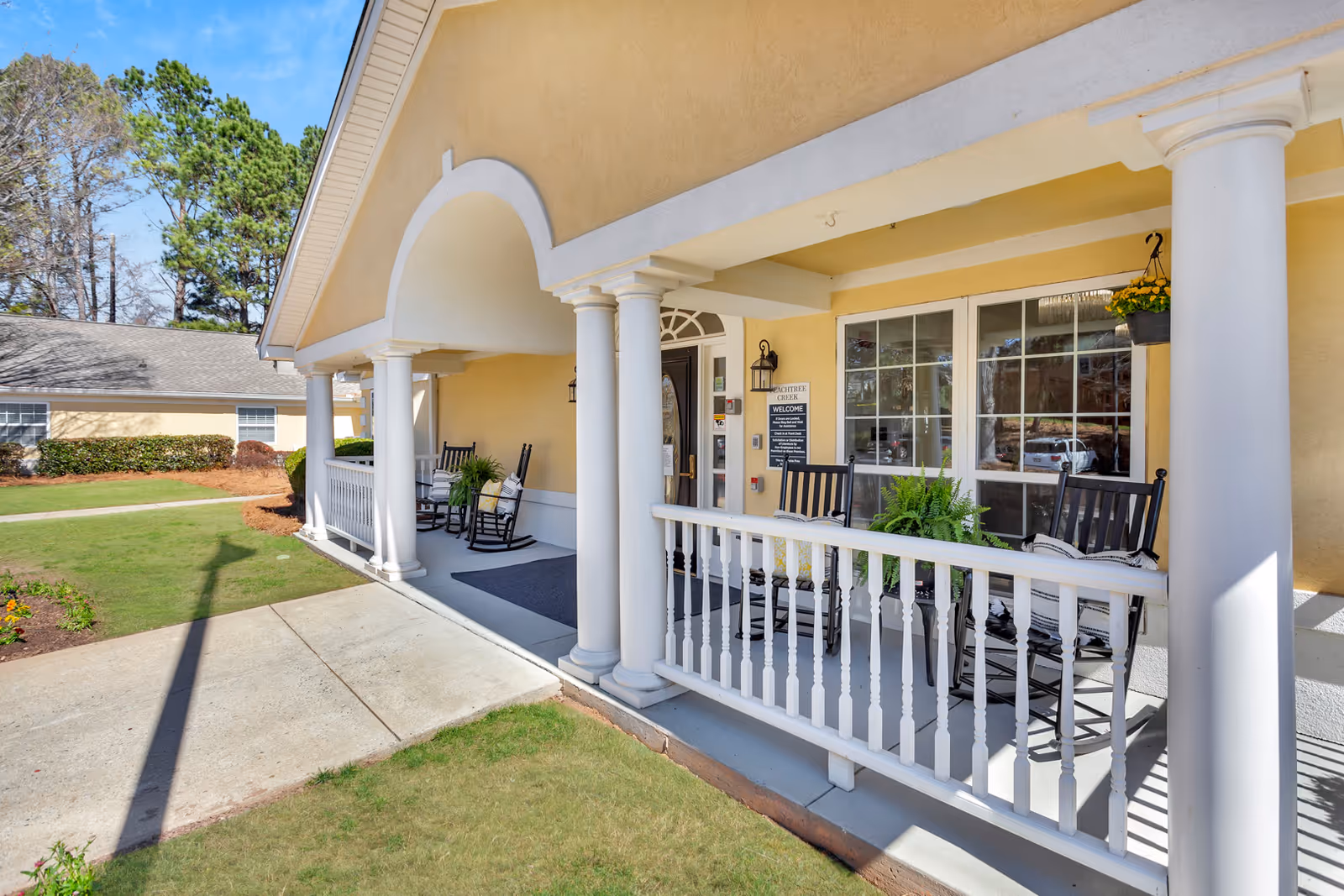 Yellow assisted living facility entrance with white columns and rocking chairs on a porch.