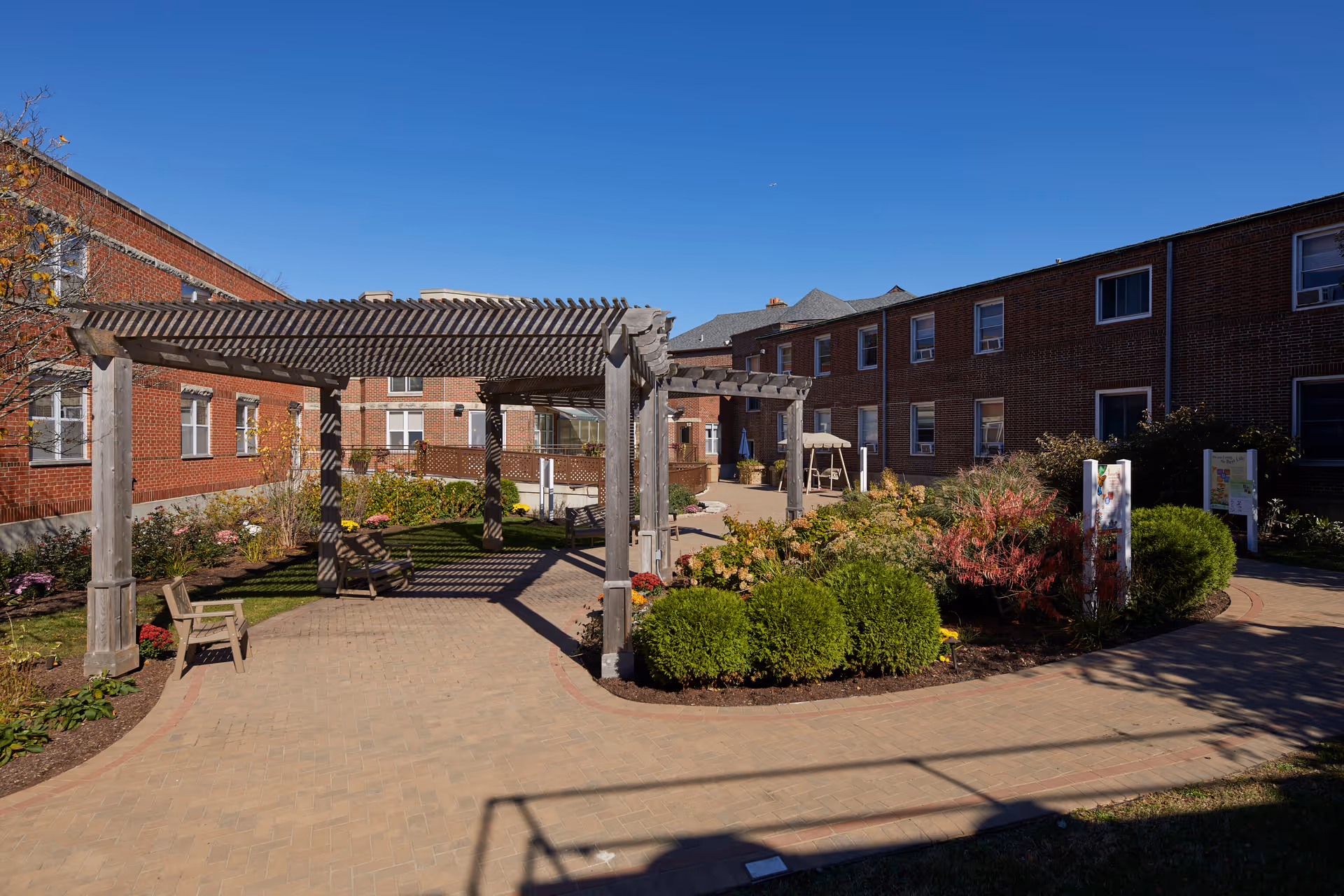 Courtyard of a senior living facility with a wooden pergola, benches, paved walkways and landscaped beds between brick buildings under a clear blue sky.