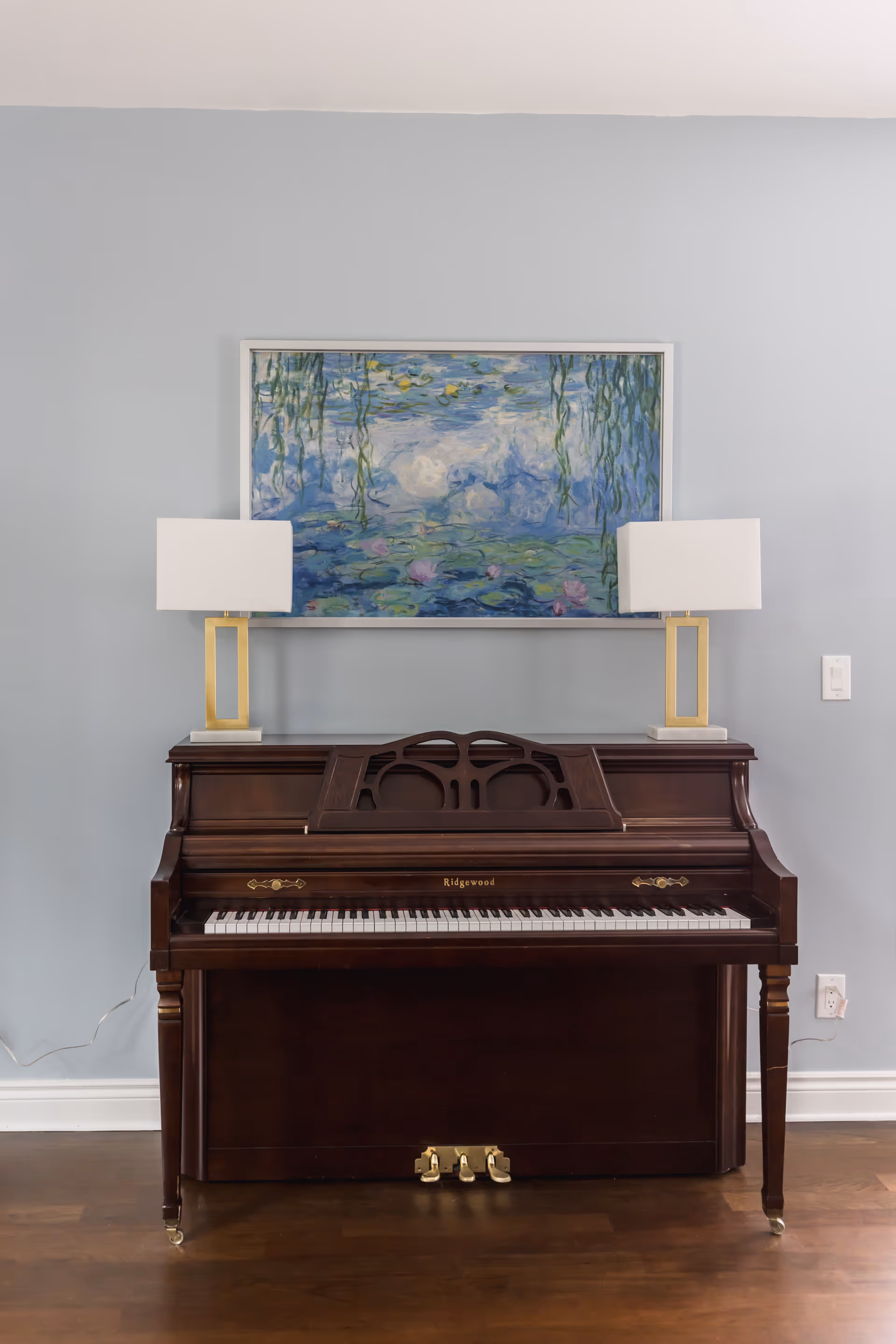 Dark wood upright piano with two lamps and a blue painting on a pale blue wall.