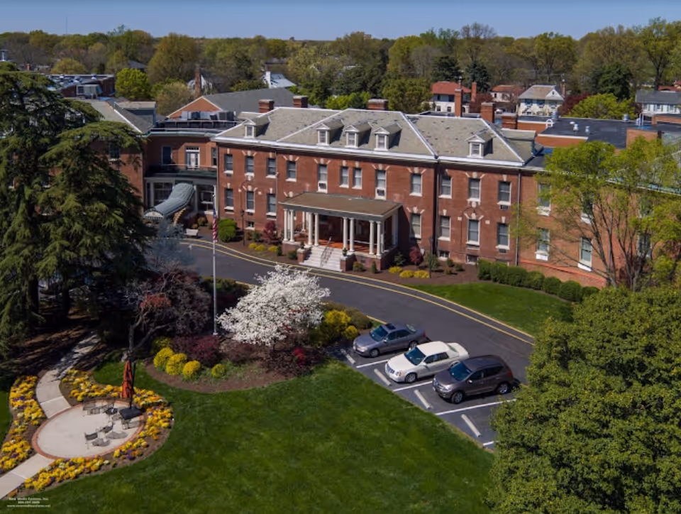 Aerial view of Hermitage Richmond, a large brick senior living facility surrounded by trees and greenery. The building has multiple windows and a covered entrance with columns. In front of the building is a circular driveway with parked cars, landscaped gardens with colorful flowers, and a small patio area with chairs and an umbrella.