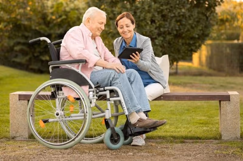 An elderly man in a wheelchair and a woman sitting on a bench outdoors, looking at a tablet together with greenery and trees in the background.
