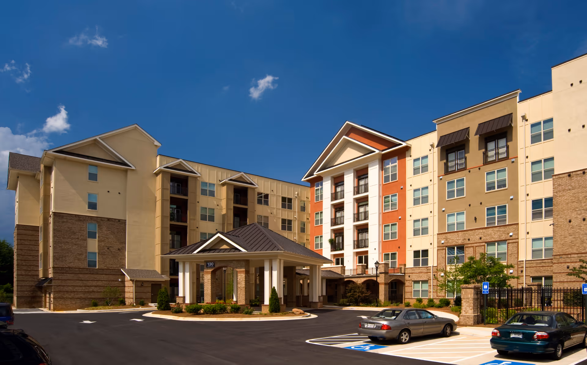 Front exterior of a multi-story senior living building with a covered entrance and adjacent parking lot under a blue sky.