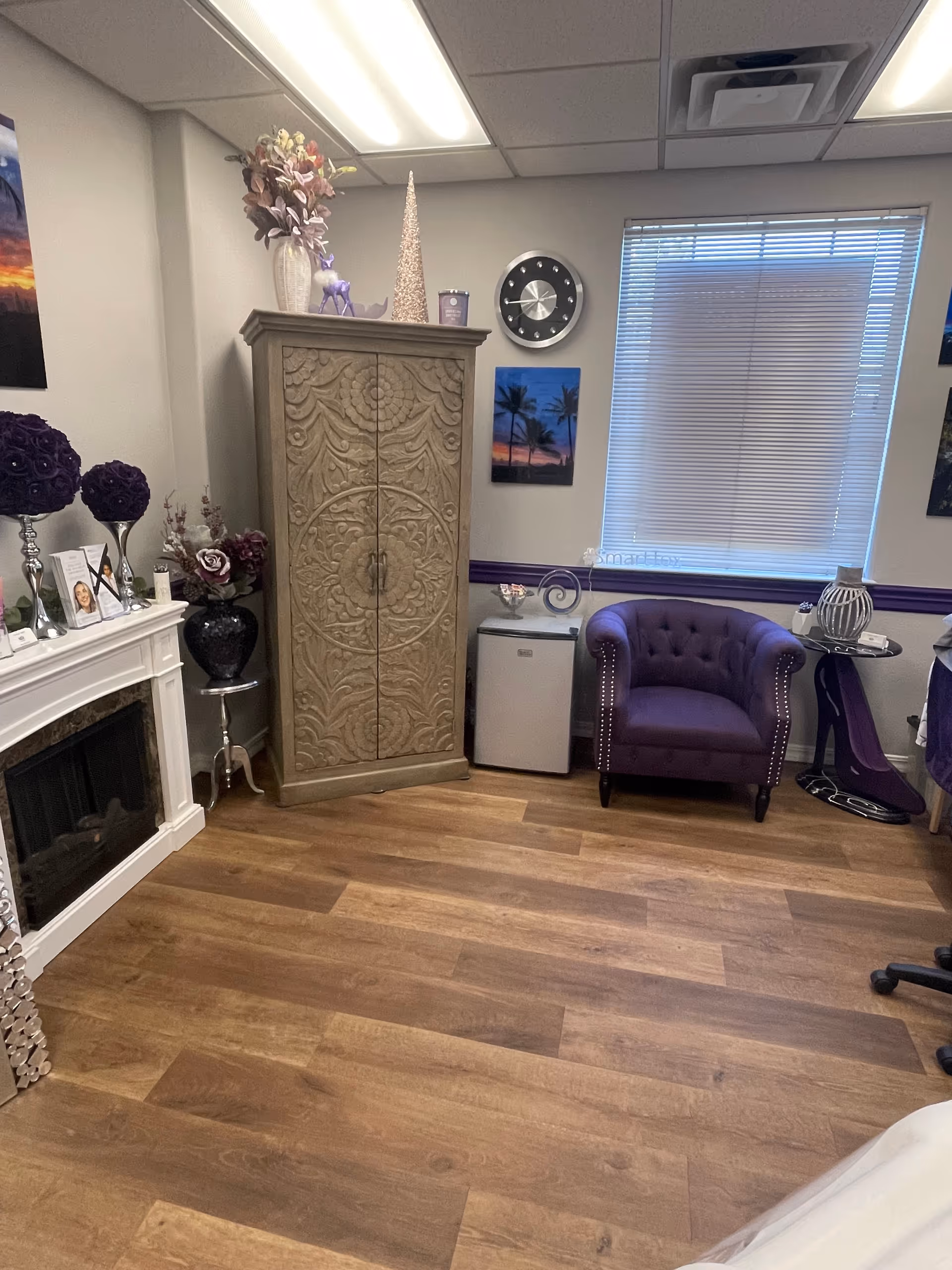 Decorative senior living lounge with purple chair, carved cabinet, fireplace, and window blinds.