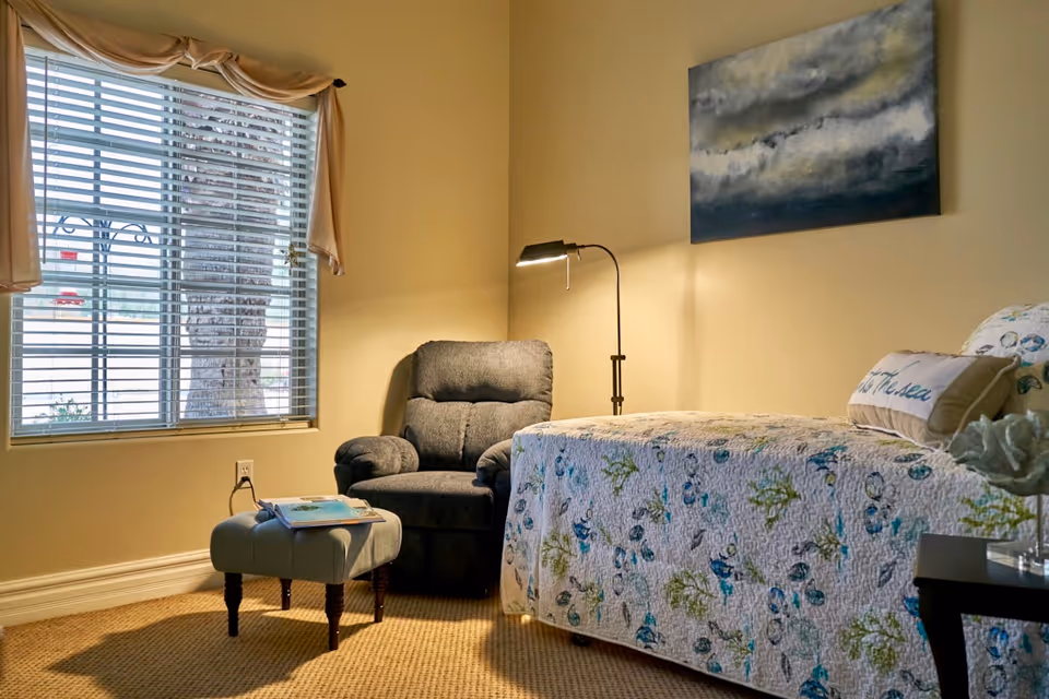 Cozy bedroom scene with a floral bed, recliner, lamp, and window blinds.
