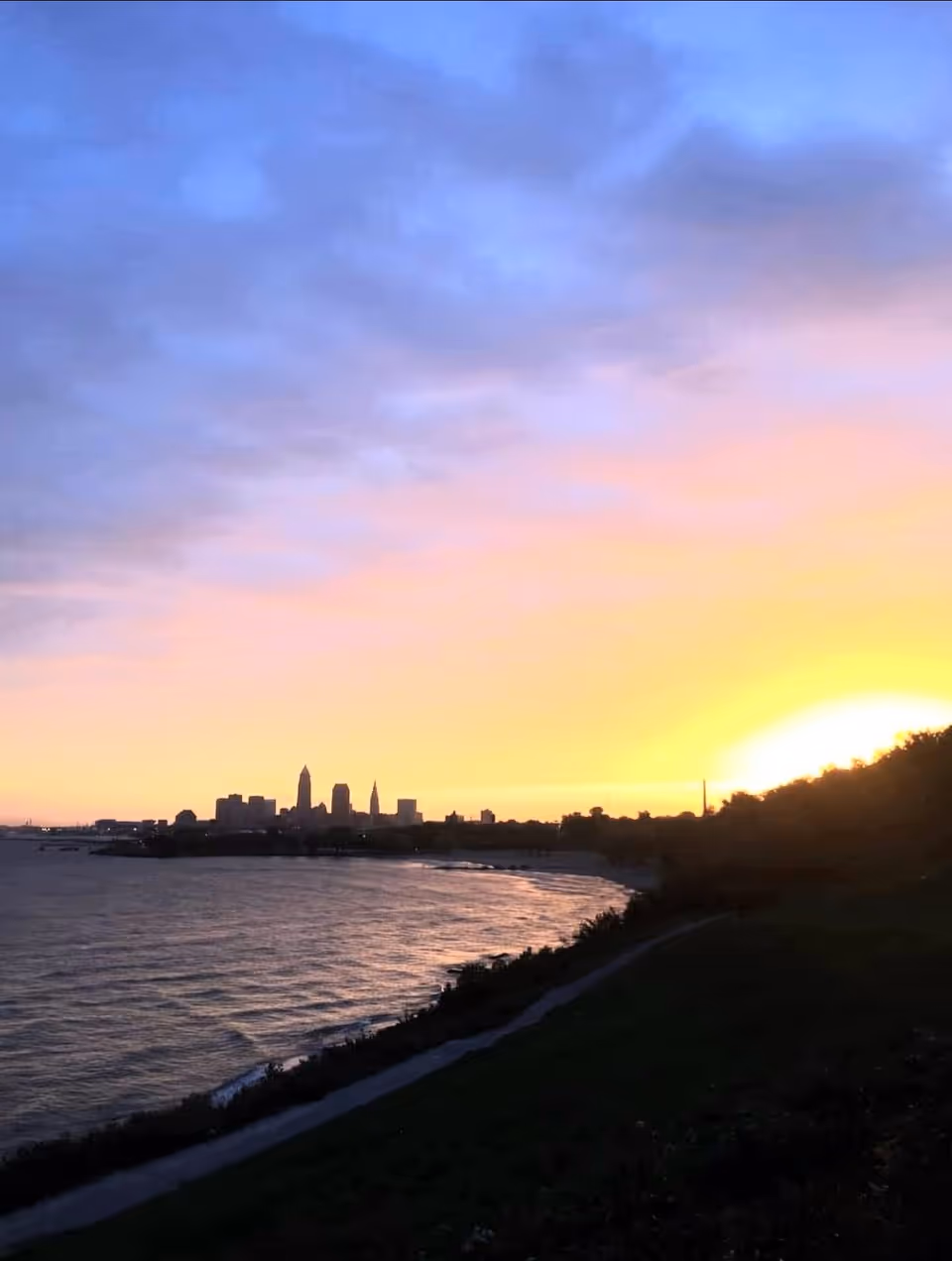 Sunset over a waterfront with a distant city skyline and a grassy path.