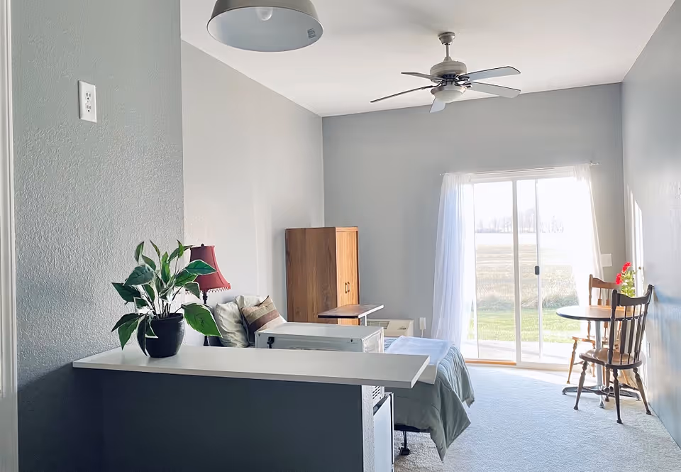 Interior senior living studio with bed, dresser, plant, dining table, and sliding patio door.