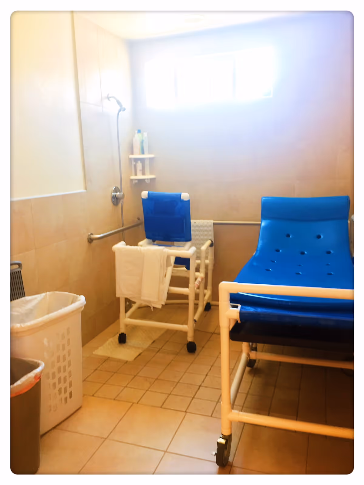 Accessible bathroom with a blue shower chair, grab bars, and a laundry hamper.