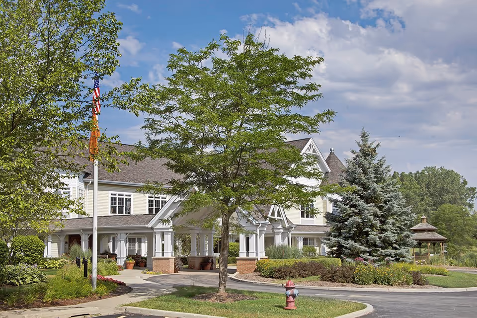 Front entrance of a senior living building with a porte-cochère, trees, flagpole, landscaped grounds and a gazebo.