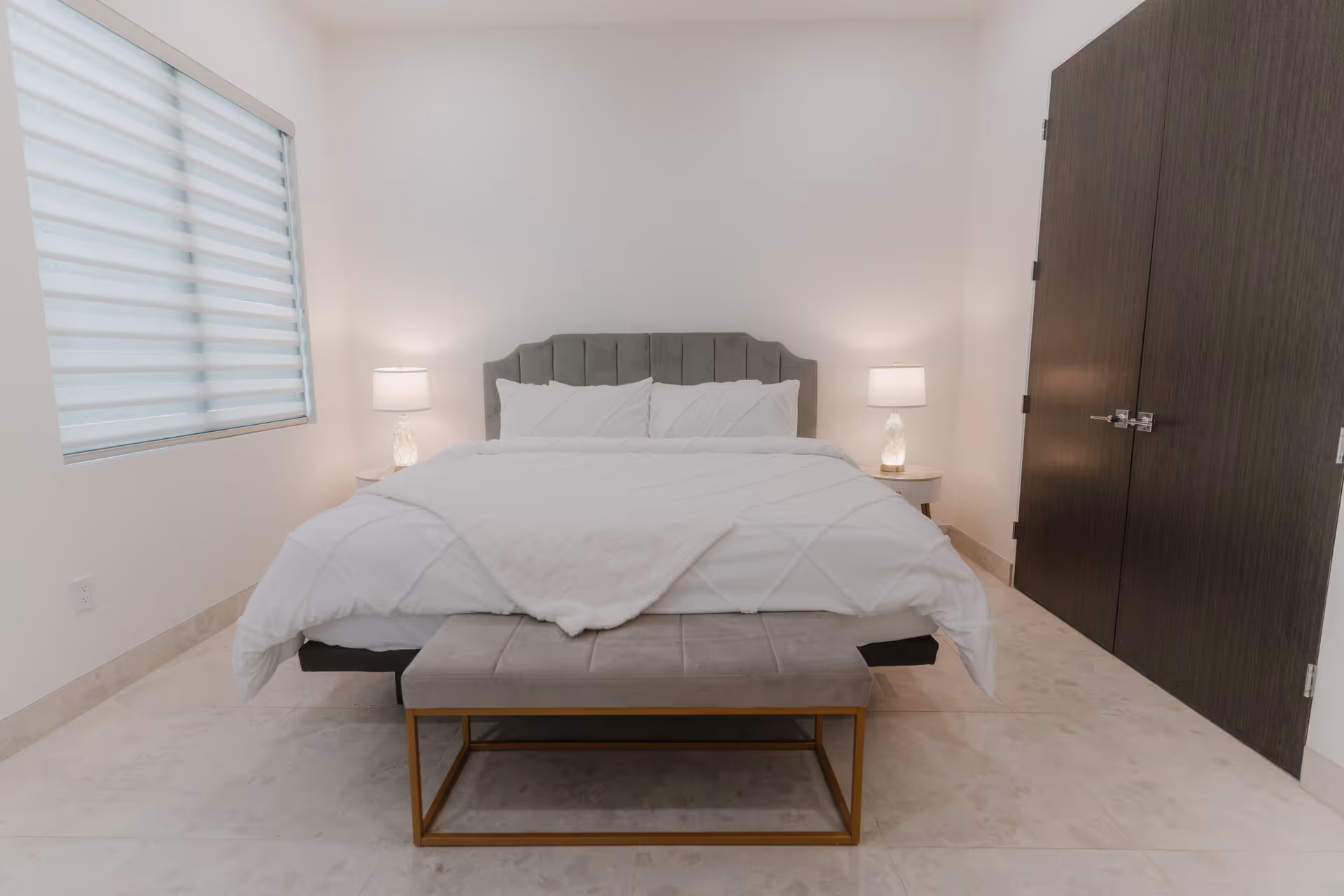 Bright bedroom in a senior facility with a bed, nightstands, lamps, window blinds, and bench.
