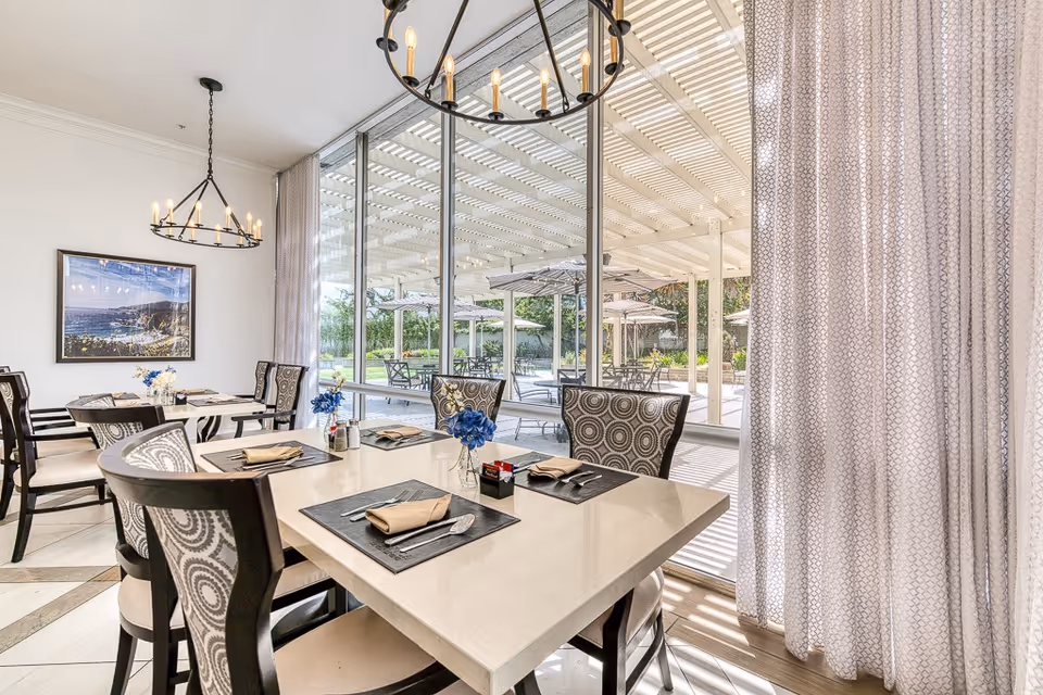 A bright dining room with a large window looking out onto an outdoor patio area with tables and umbrellas. The dining room has a white table set with placemats, napkins, and cutlery, surrounded by chairs with patterned backs. Two chandeliers hang from the ceiling, and a framed picture is on the wall.