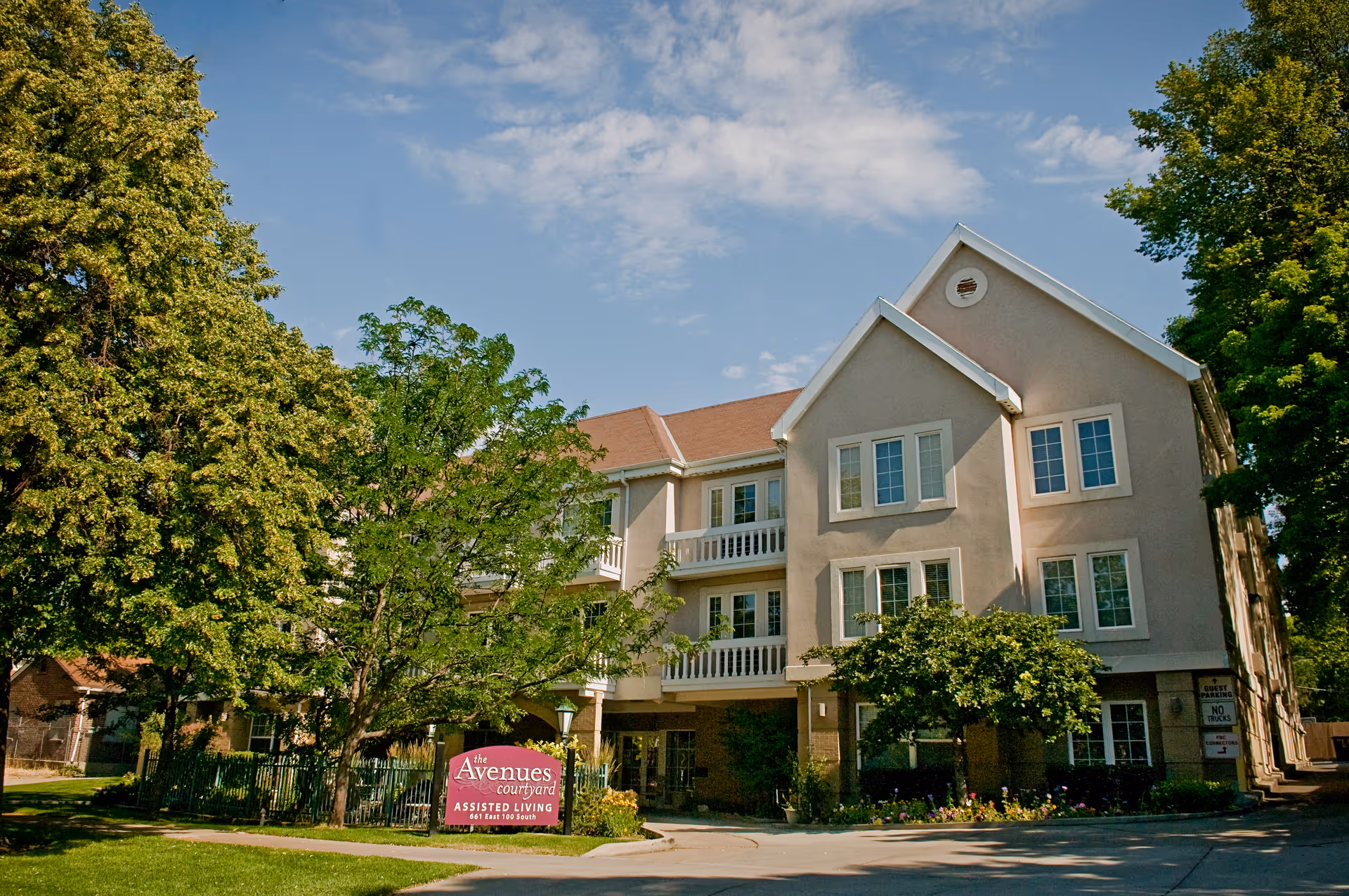 Three-story beige assisted living building with balconies, trees, and a pink entrance sign.