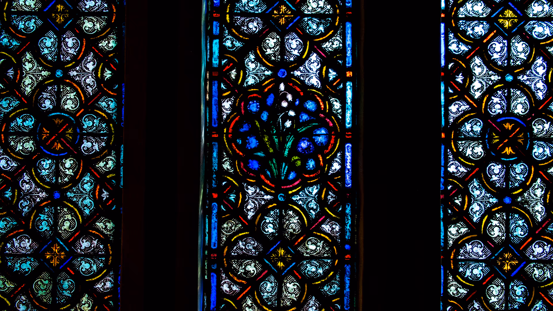 Close-up of three vertical stained glass windows with intricate floral and geometric patterns in blue, green, yellow, and red colors.