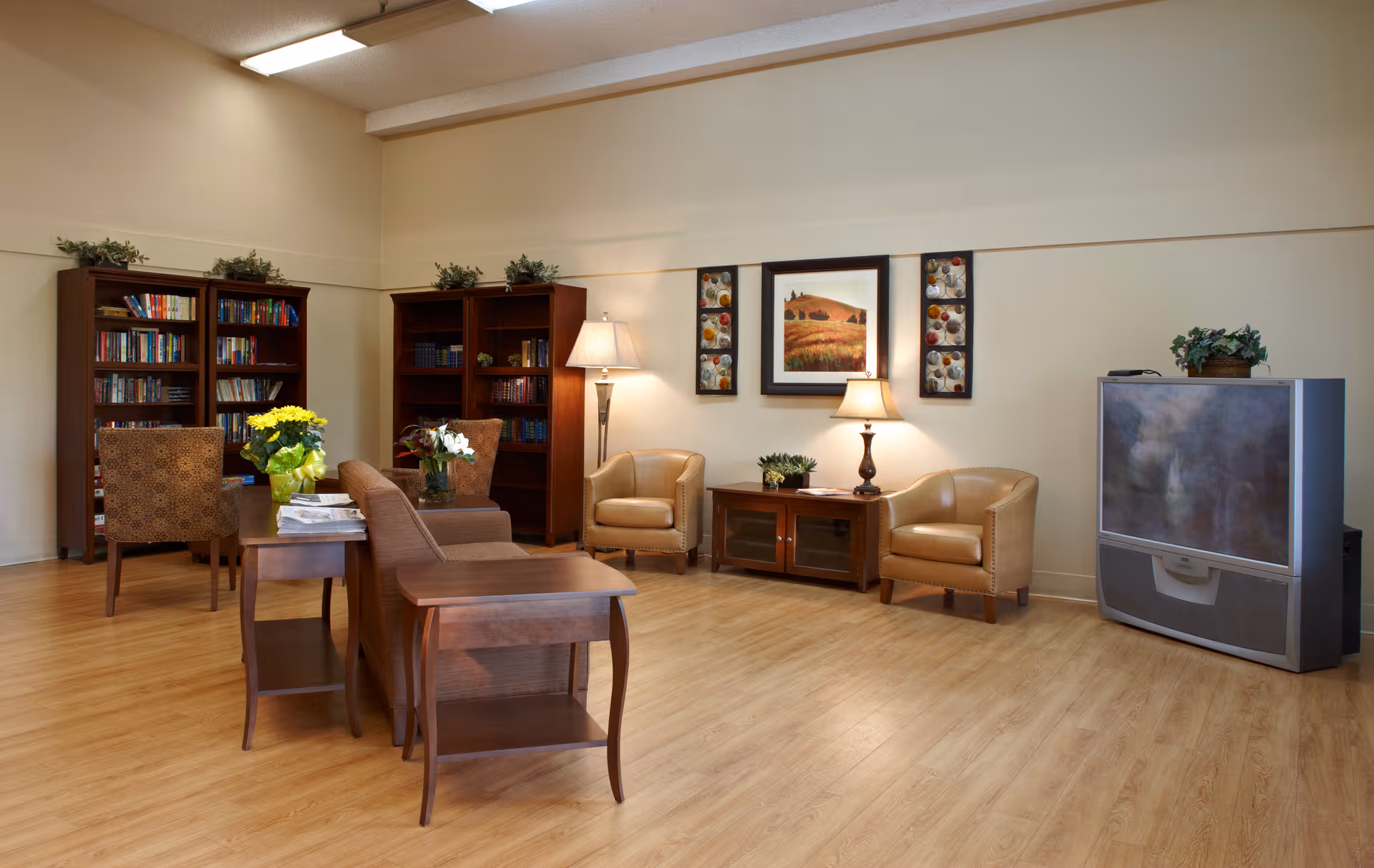 Senior living lounge with bookshelves, seating, lamps, and a large television.