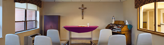 A small chapel room with a table covered by a purple cloth, a crucifix on the wall, an organ, and several chairs arranged facing the table. There are windows on both sides letting in natural light.