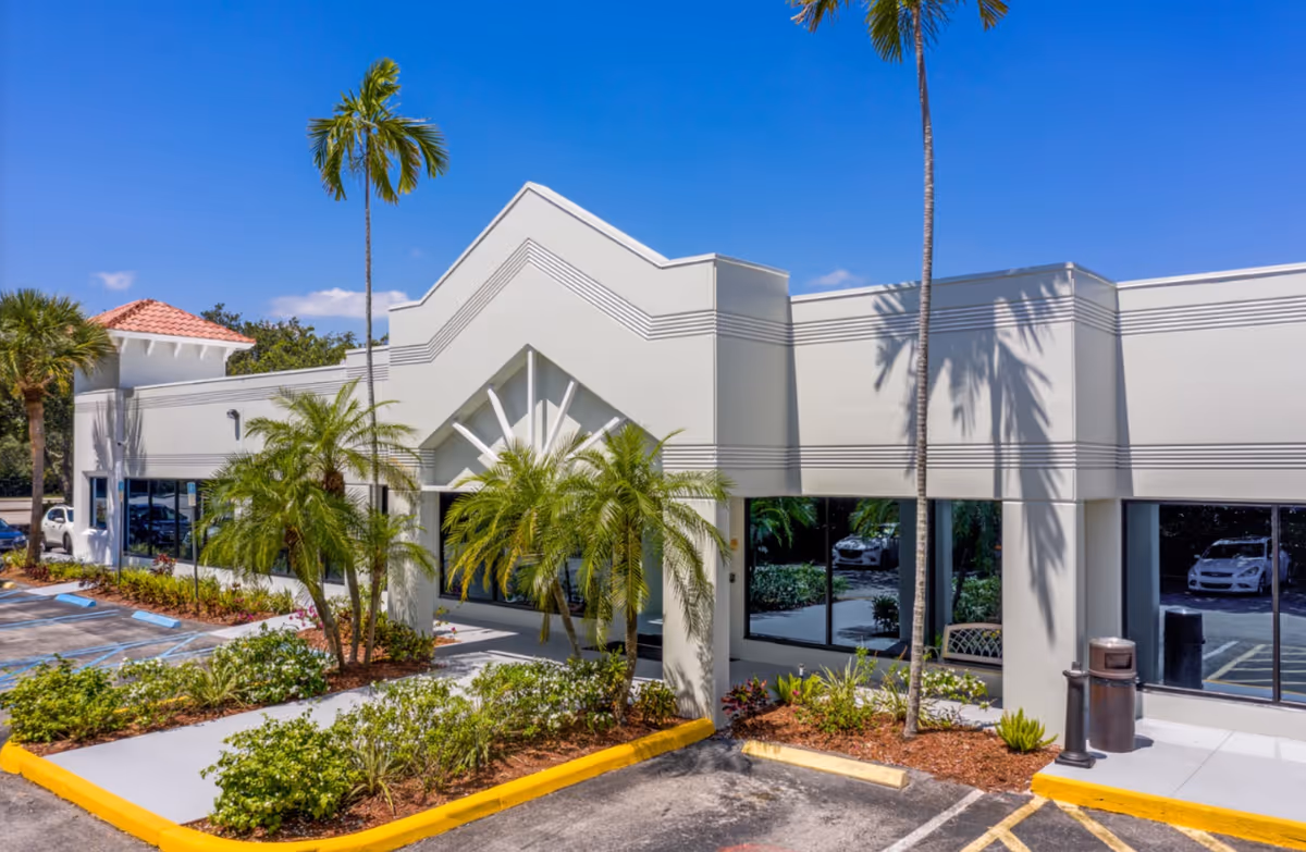 Modern beige senior living facility exterior with palm trees, glass windows, and a parking lot.