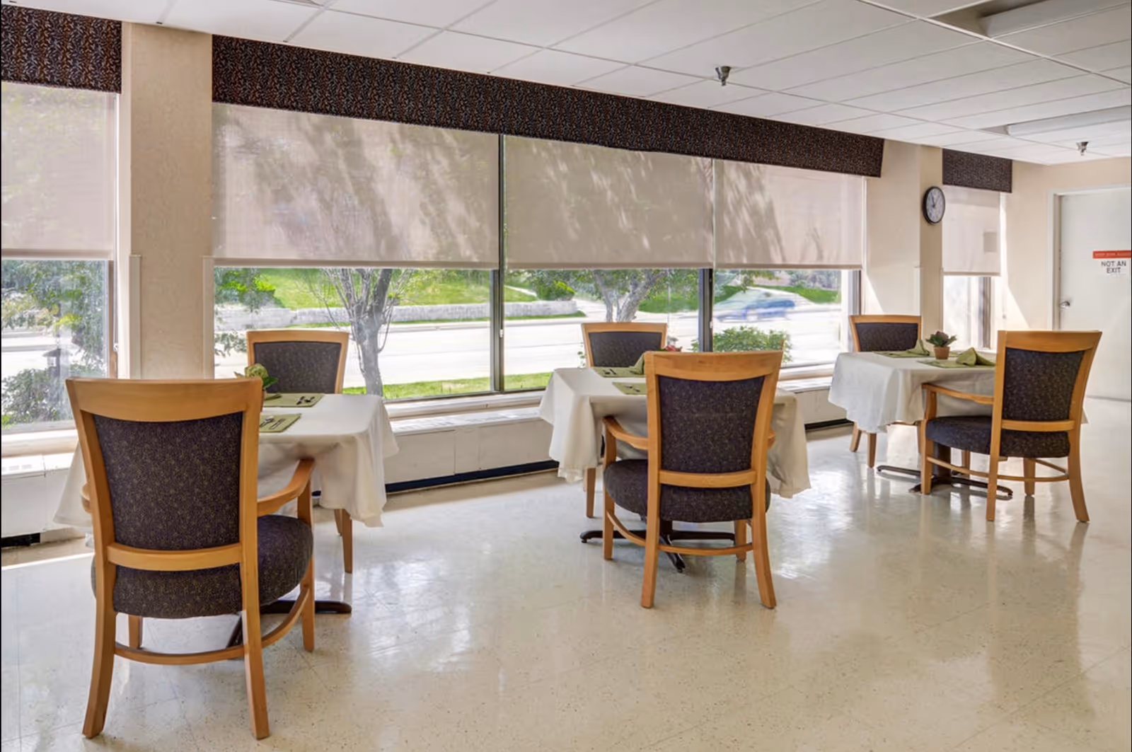 Dining room with white-tablecloth tables and wooden chairs along large windows.