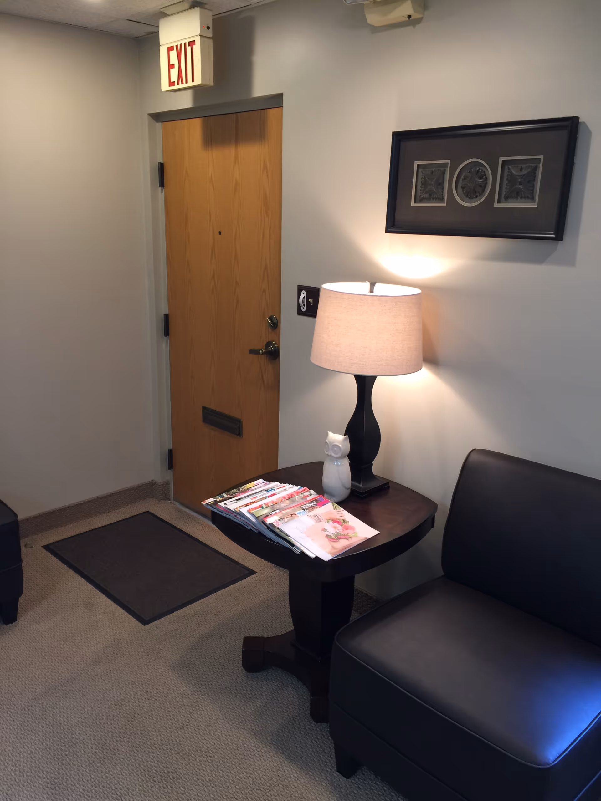 Indoor waiting area with exit sign, wooden door, lamp, magazines, and sofa.