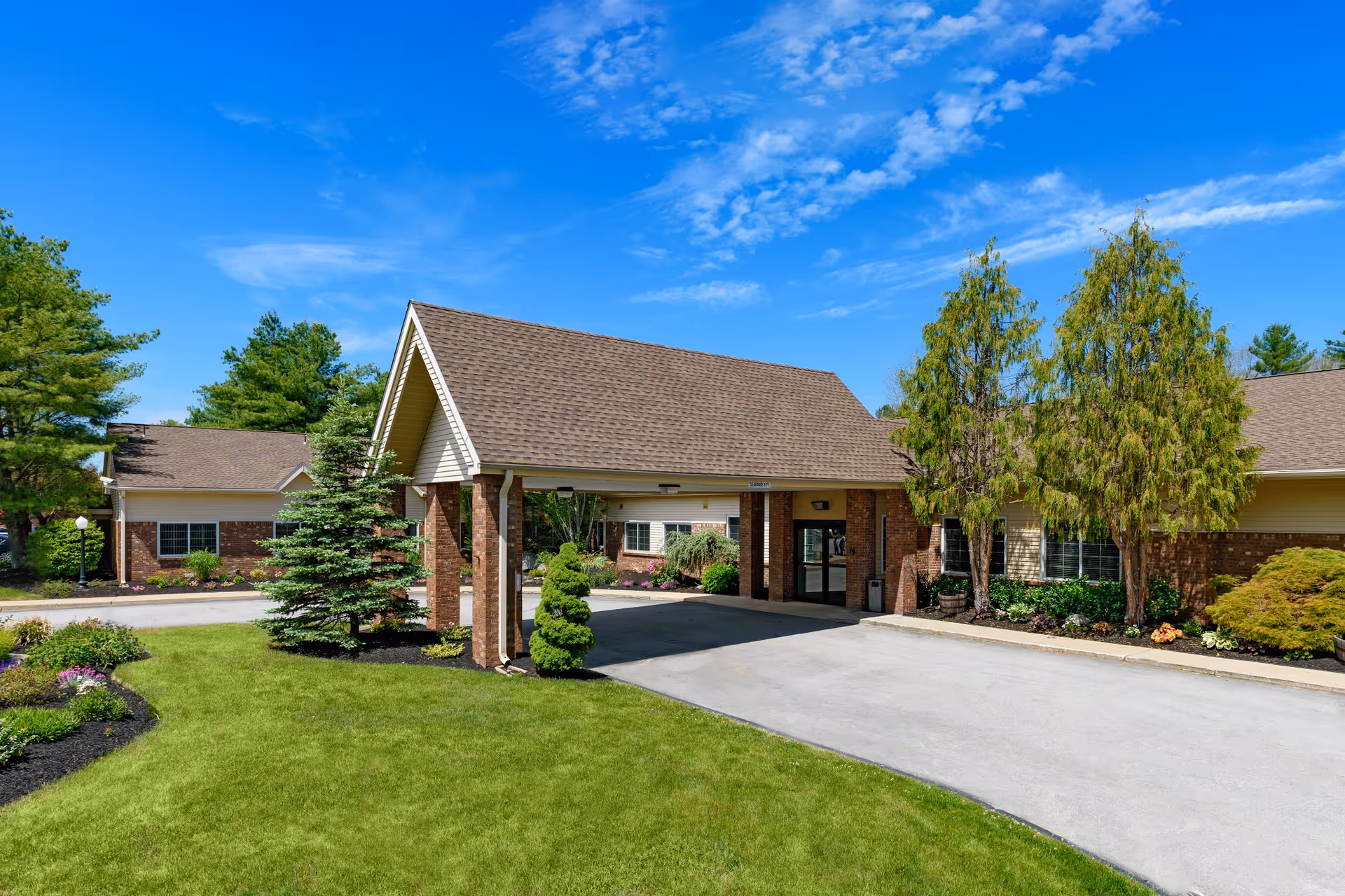 Exterior view of The Oaks senior living facility entrance with a covered drop-off area, surrounded by well-maintained landscaping including trees, shrubs, and green grass under a bright blue sky with scattered clouds.