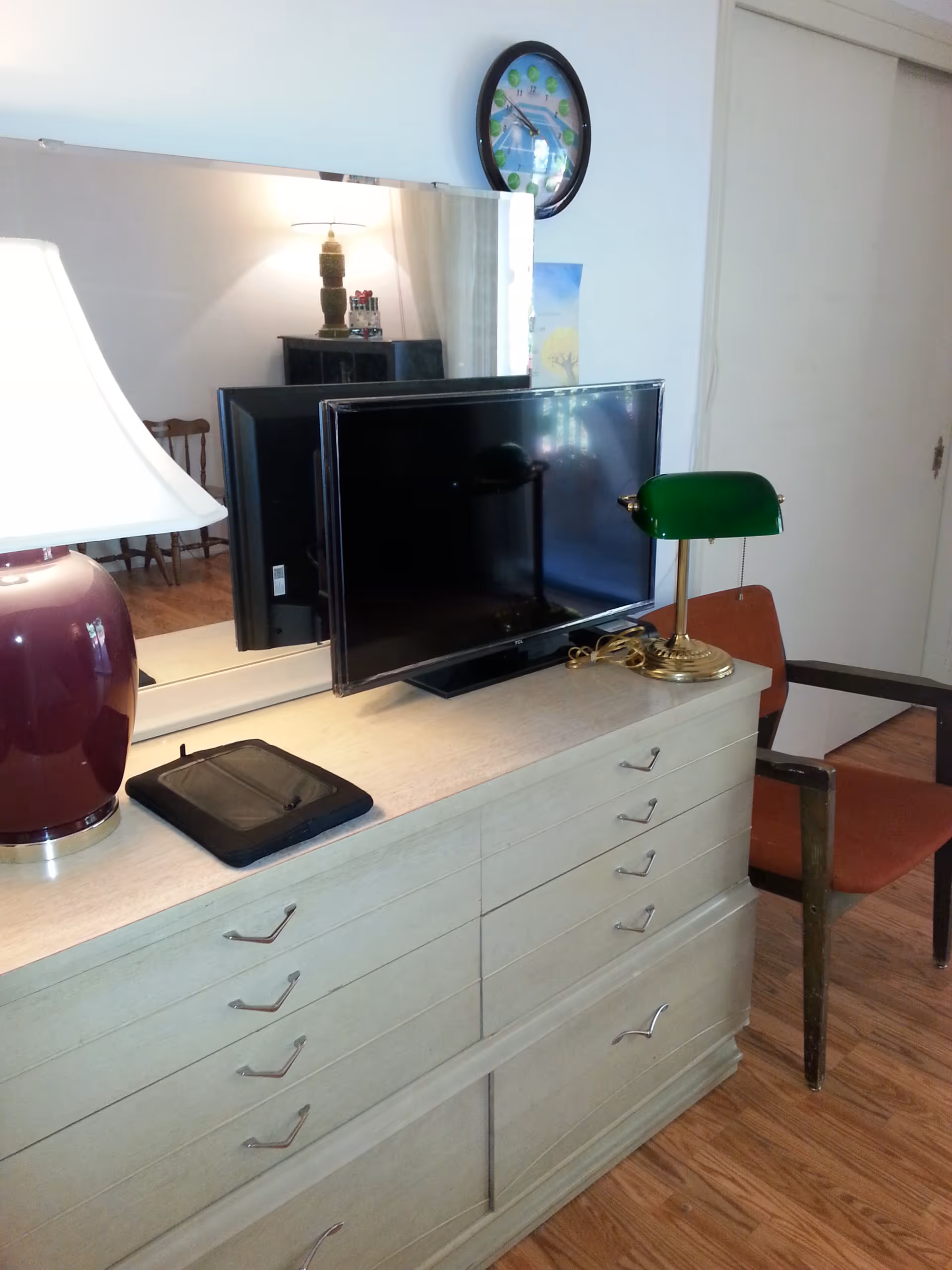 A dresser with multiple drawers topped with a flat-screen TV, a green banker’s lamp, a tablet, and a large purple lamp. Behind the dresser is a large mirror reflecting part of the room, including a wooden chair and a lit lamp on a cabinet. A round wall clock with a blue and green design hangs above the TV. The floor is wooden, and a red cushioned chair with wooden arms is positioned next to the dresser.