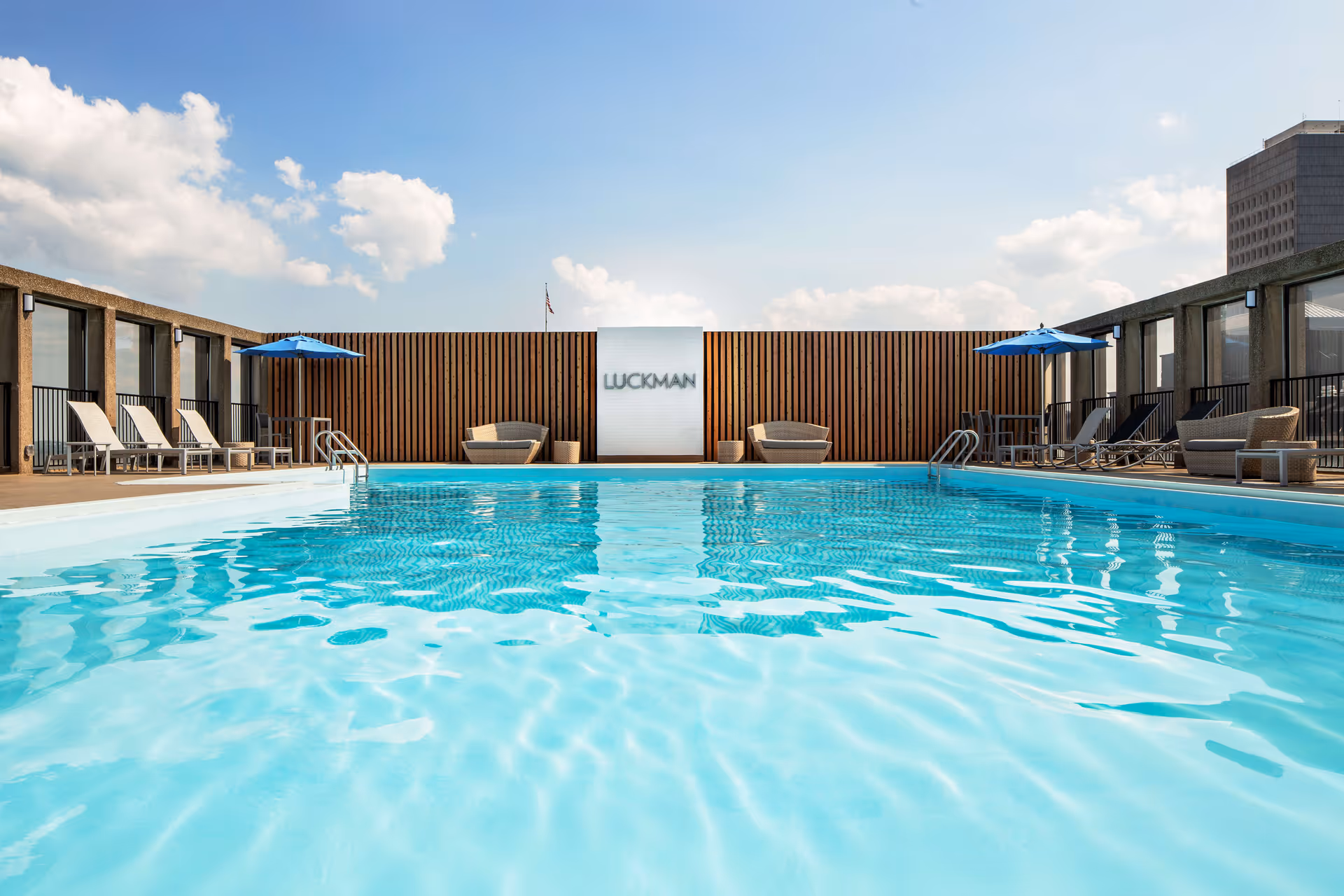Outdoor swimming pool with clear blue water surrounded by lounge chairs, umbrellas, and seating areas on a rooftop with a wooden privacy fence and a sign that reads 'LUCKMAN'. The sky is partly cloudy.