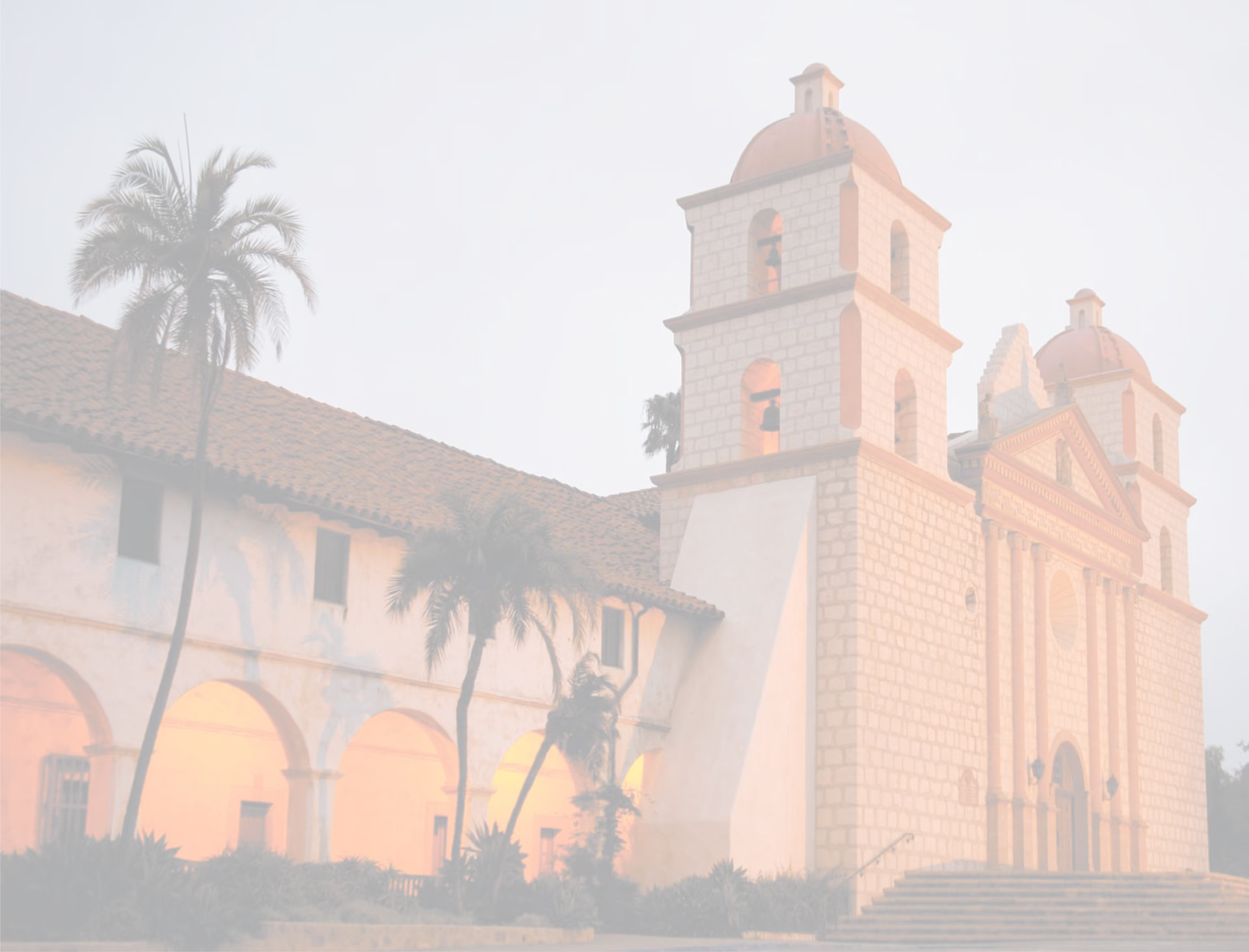 Mission-style front exterior of a stone building with bell towers and arched entryway.