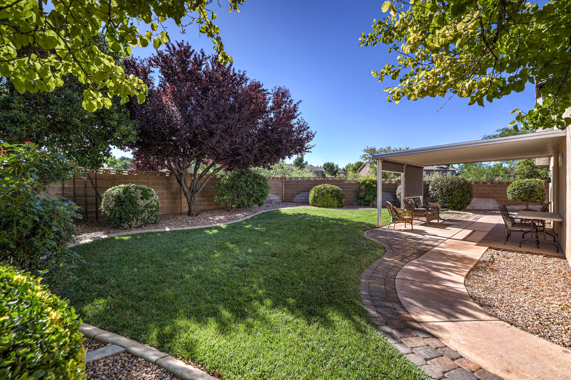 A well-maintained outdoor garden area with a green lawn, various bushes, and a tree with dark red leaves. There is a paved walkway curving through the garden leading to a covered patio area with outdoor chairs and a table. The sky is clear and blue.