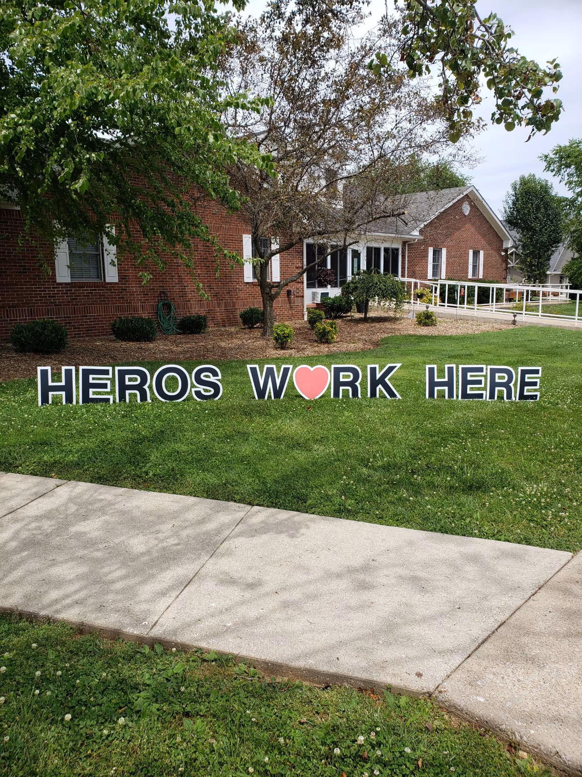 A grassy lawn in front of a brick building with white window shutters and a ramp. On the lawn, large black and white letters spell out 'HEROS W❤️RK HERE' with a red heart symbol replacing the letter 'O' in the word 'WORK'. Trees and shrubs are visible around the building.