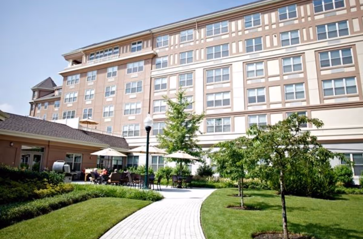 Well-maintained courtyard with a paved walkway, patio tables and umbrellas in front of a multi-story senior living building.