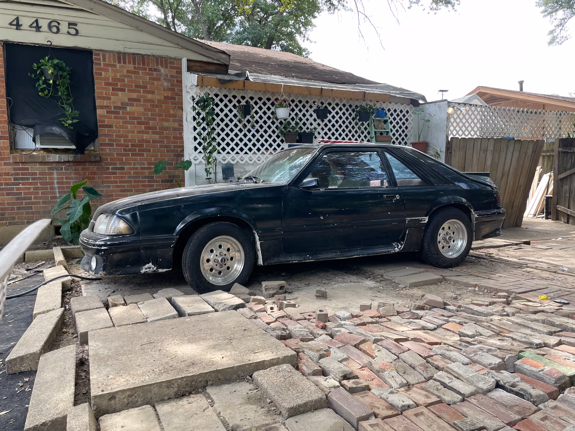 Black car parked on a brick driveway beside a lattice-front house.
