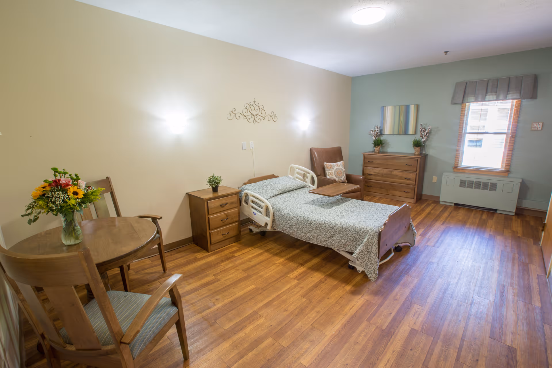 A senior living facility bedroom with a single hospital-style bed covered with a patterned bedspread, a wooden nightstand with a small plant, a brown armchair with a decorative pillow, a wooden dresser with two plants and a colorful painting above it, a window with a valance, and a small round wooden table with two chairs and a vase of flowers. The room has wood flooring and soft wall lighting.