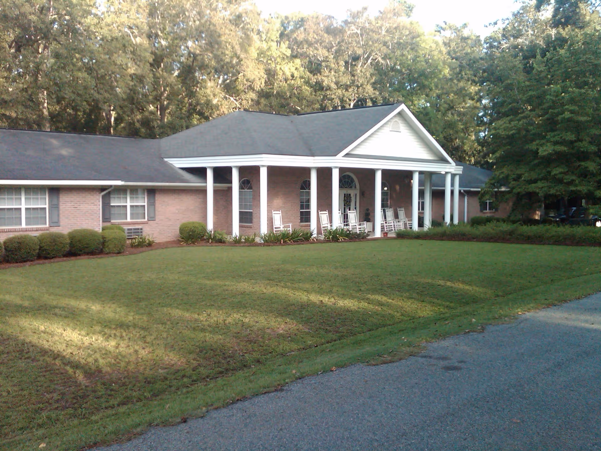 Single-story brick building with a covered front porch supported by white columns and rocking chairs, facing a manicured lawn and driveway.