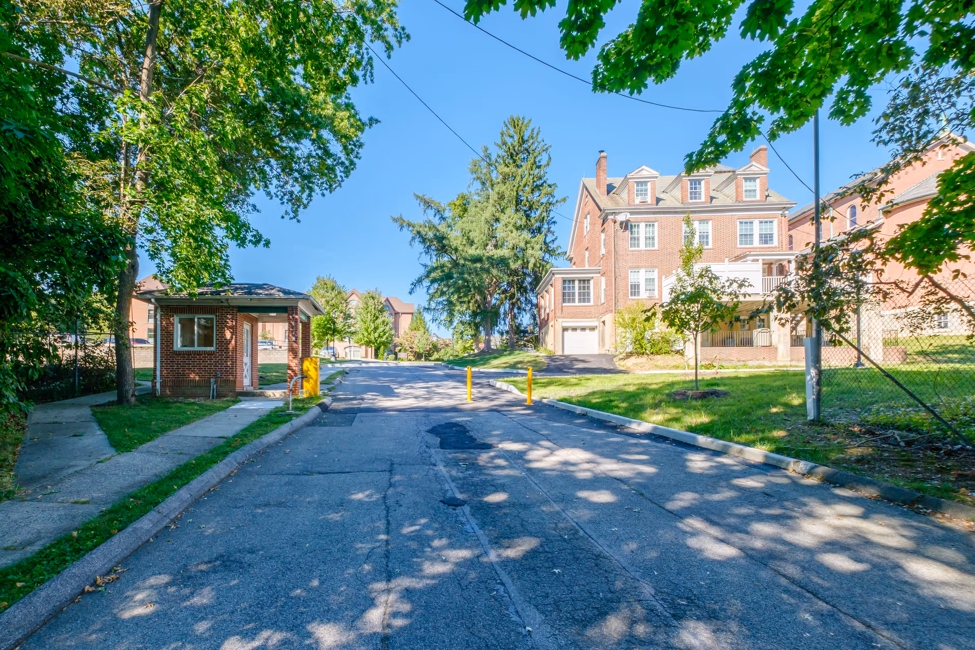 A paved driveway leading uphill with a small brick guardhouse on the left and a large multi-story brick building on the right, surrounded by green trees and grass under a clear blue sky.