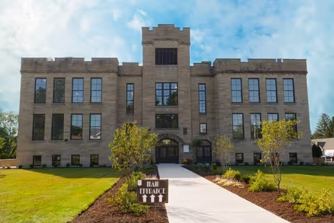 A large stone building with doorway, symmetrical windows, and a paved walkway leading to the entrance.