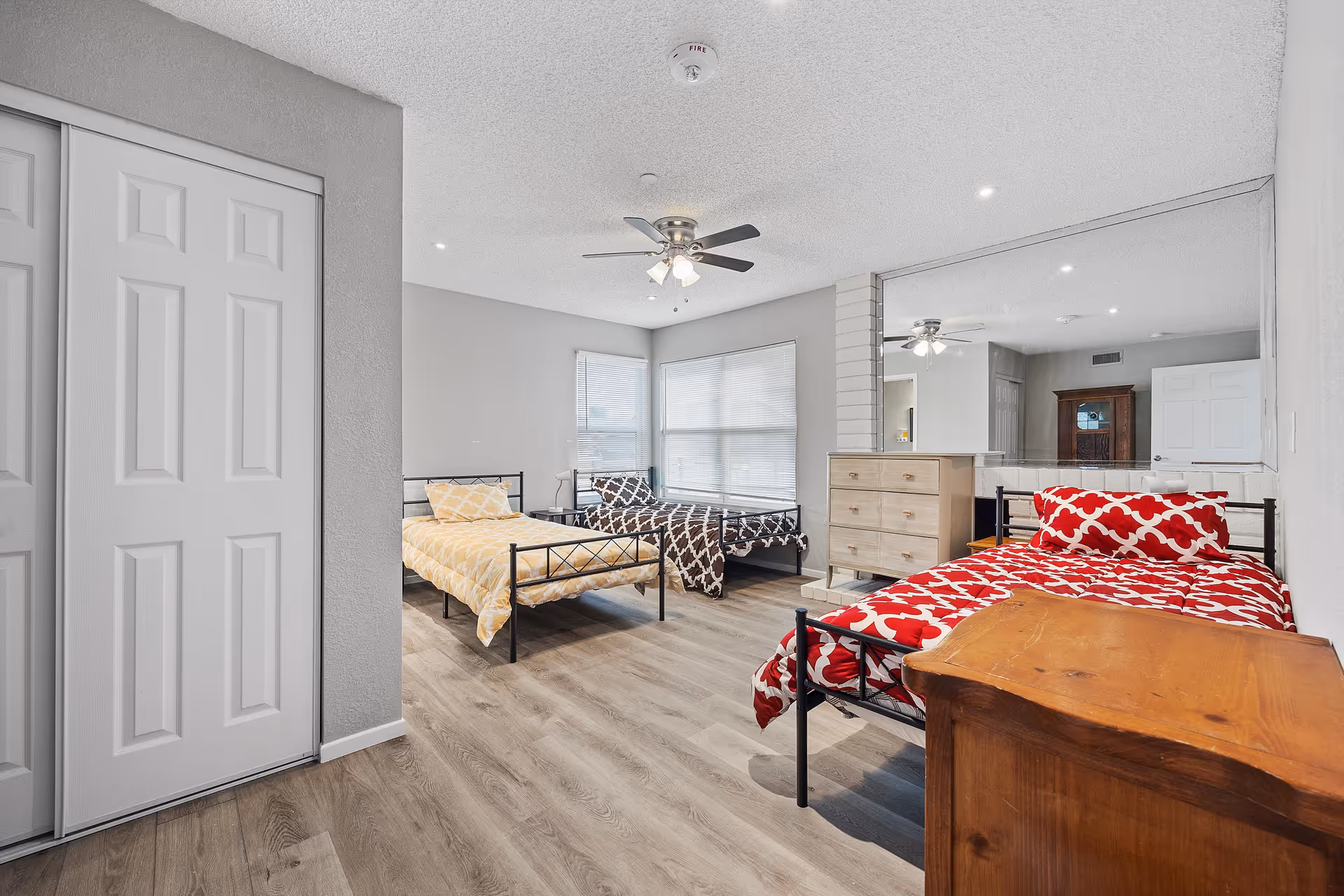 A shared bedroom with three metal-frame beds, dresser, and mirrored wall in a bright facility.