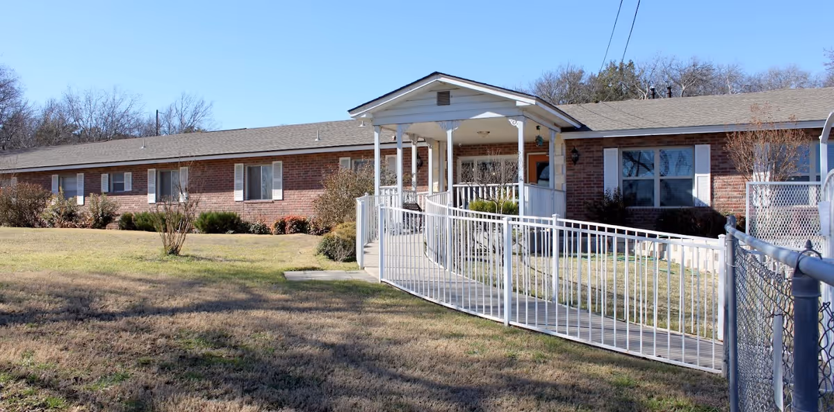 Single-story brick assisted living building with a covered entrance and a white wheelchair ramp across a grassy front lawn.