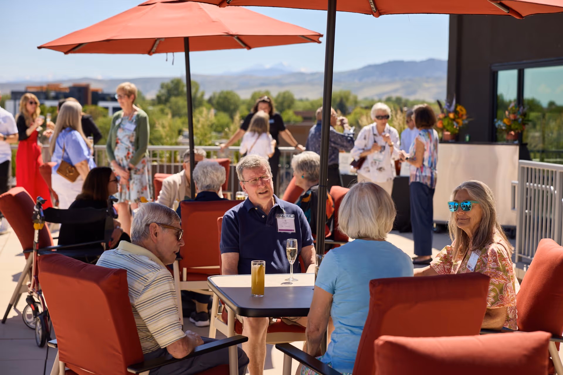 Residents chat and socialize around tables on an outdoor terrace with umbrellas and mountains in background.