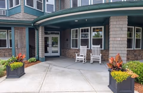 Entrance area of a senior living facility with two white rocking chairs on a covered porch. The building exterior features stone and shingle siding with large windows. Two planters with green and red foliage flank the concrete walkway leading to the door.